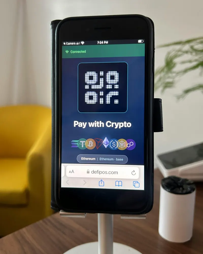 A phone on a stand at a shop counter, running the DefiPOS customer display: the LED logo, "Pay with Crypto" and the coins accepted.